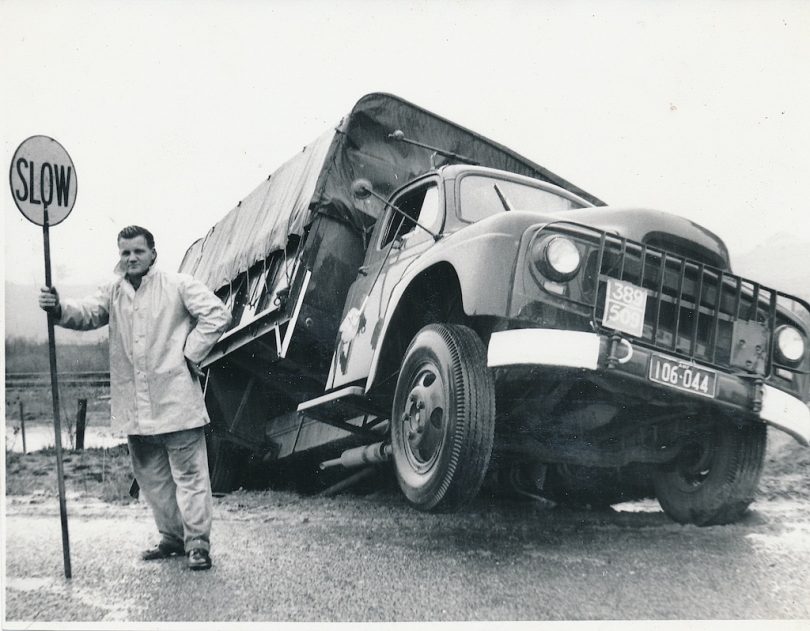 Historical image of man standing with 'slow' sign next to crashed truck on Old Hume Highway