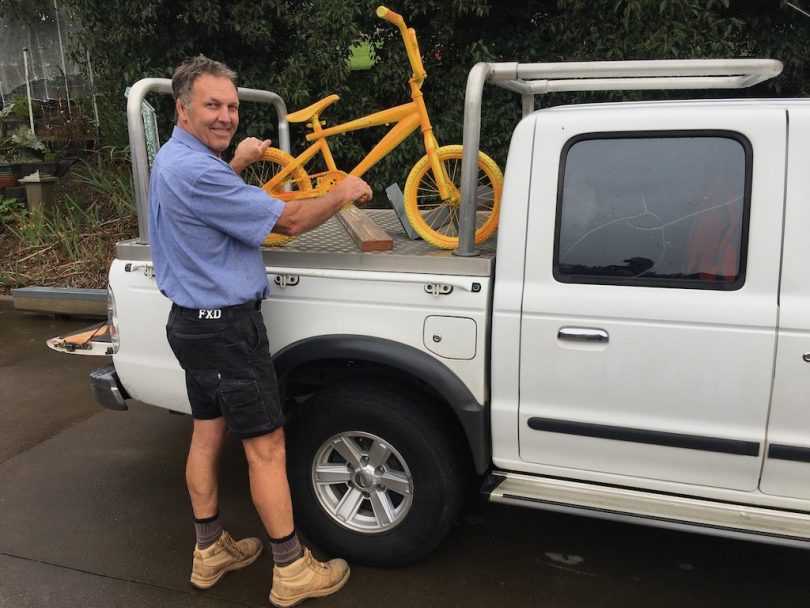 A man loads a yellow child-sized bike onto the back of his ute.