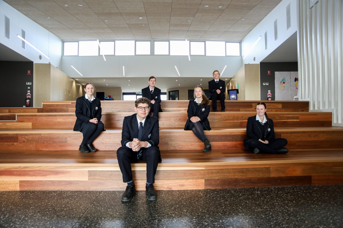 Six students sit spread out on wooden stairs inside a large room.