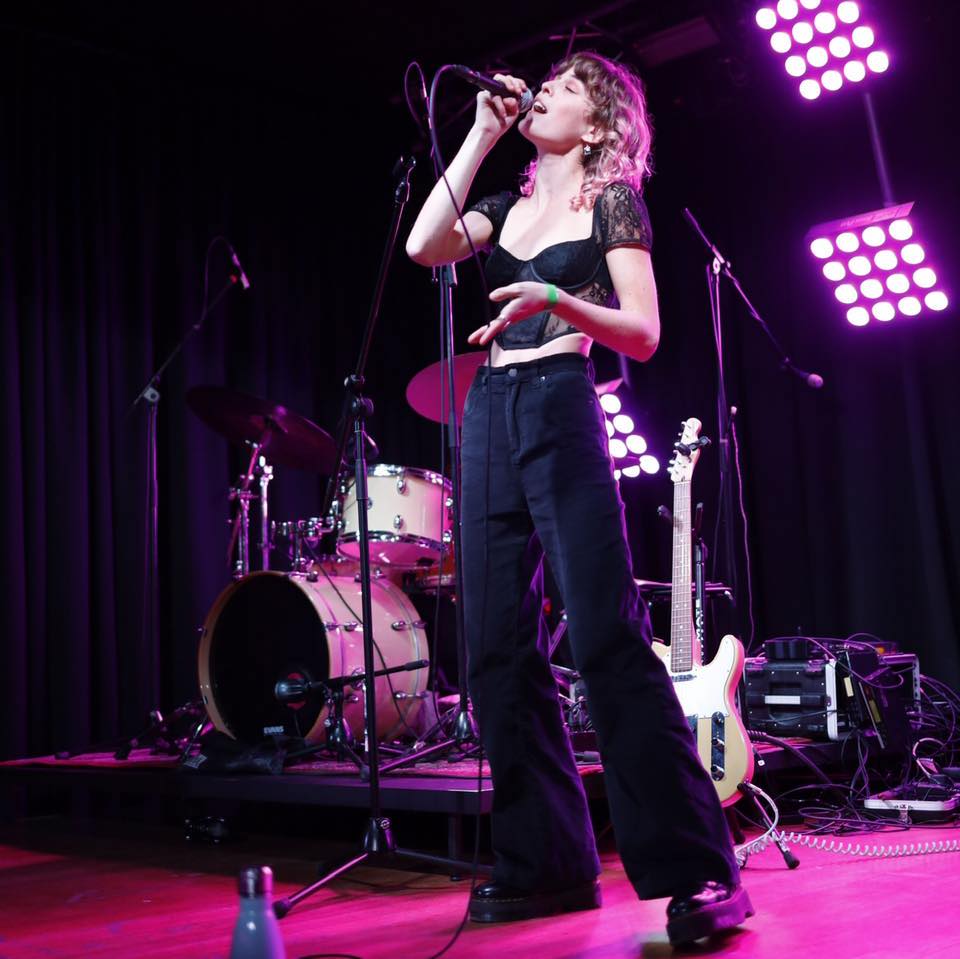 A photograph of Ava Martina singing in an all-black outfit and a microphone in one hand