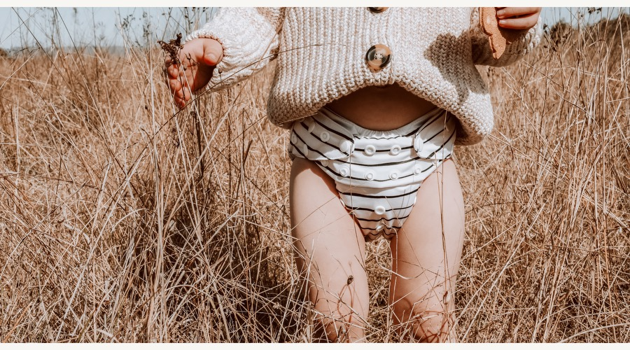 Baby wearing a beige sweater in wheat field with black and white striped reusable nappy.