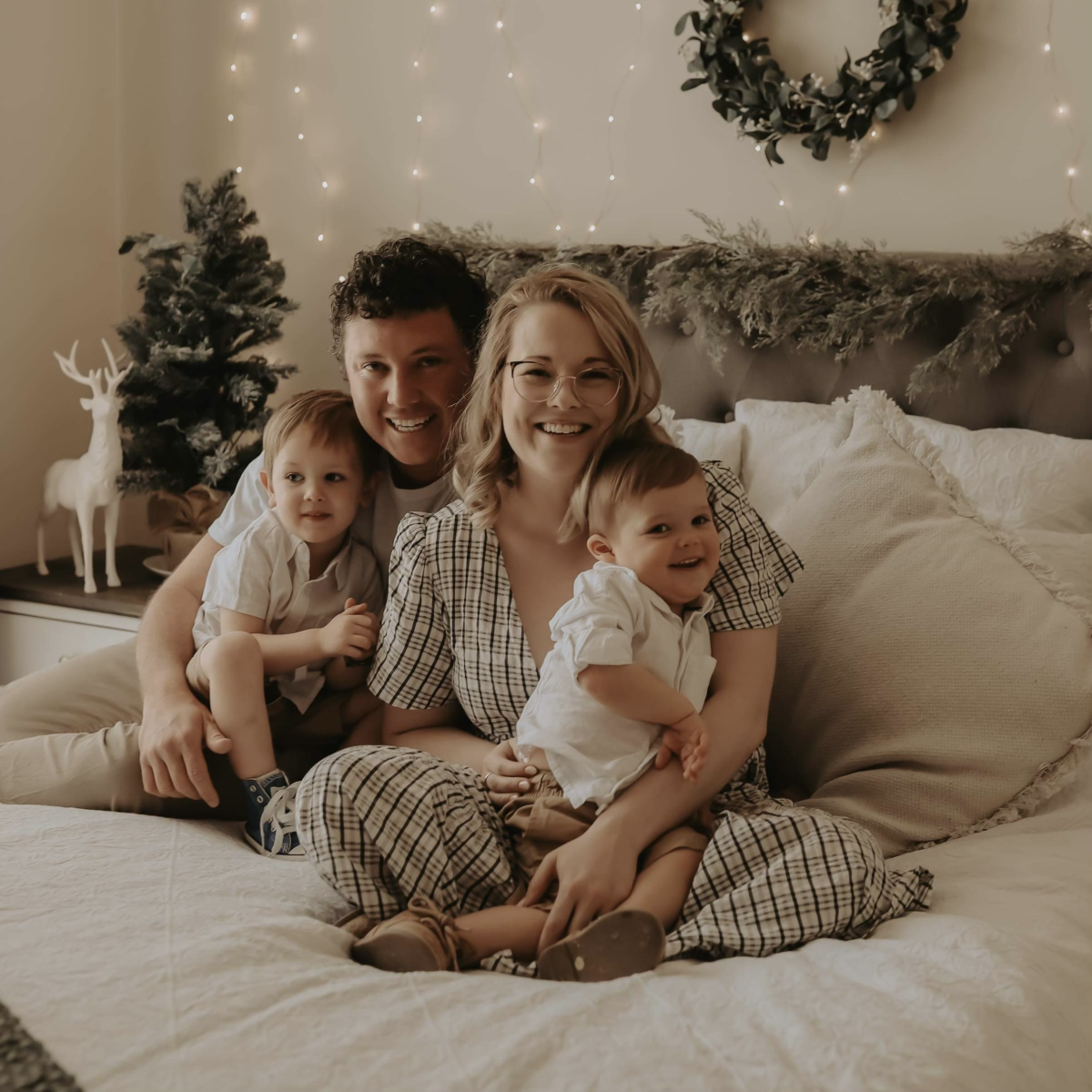 Mother, father and two sons sit on a white bed at Christmastime. 