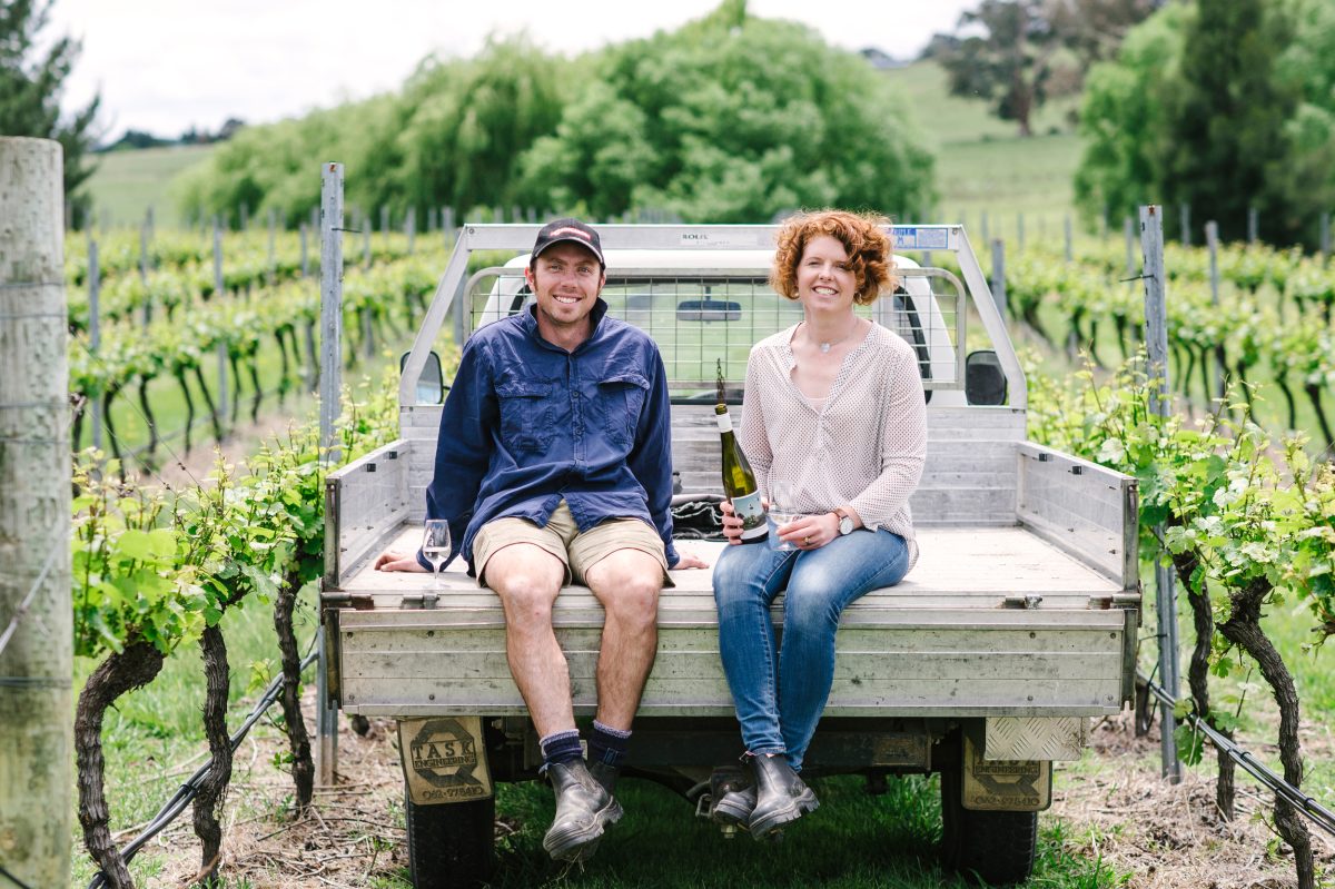 A man and woman sit on the back tray of a ute parked in a vineyard. She is holding a bottle of wine. 