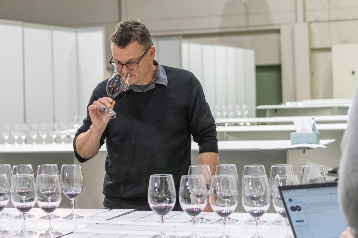 Matt Harrop sniffs a glass of wine while standing at a table set with several glasses of different wines. 
