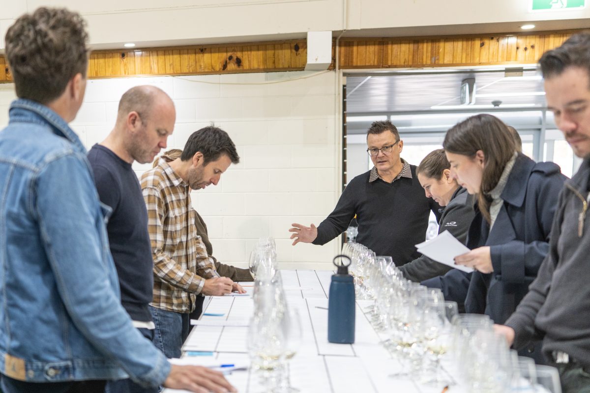 Judges stand around a table set with wine glasses, as a man speaks at the front of the room.