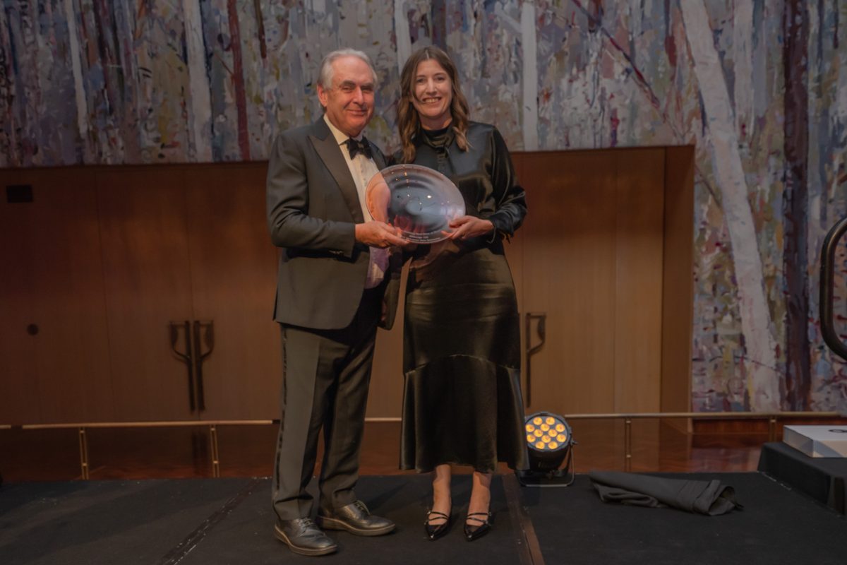 An older man in suit and bow tie presents a glass plate trophy to a woman in formal black satin dress.