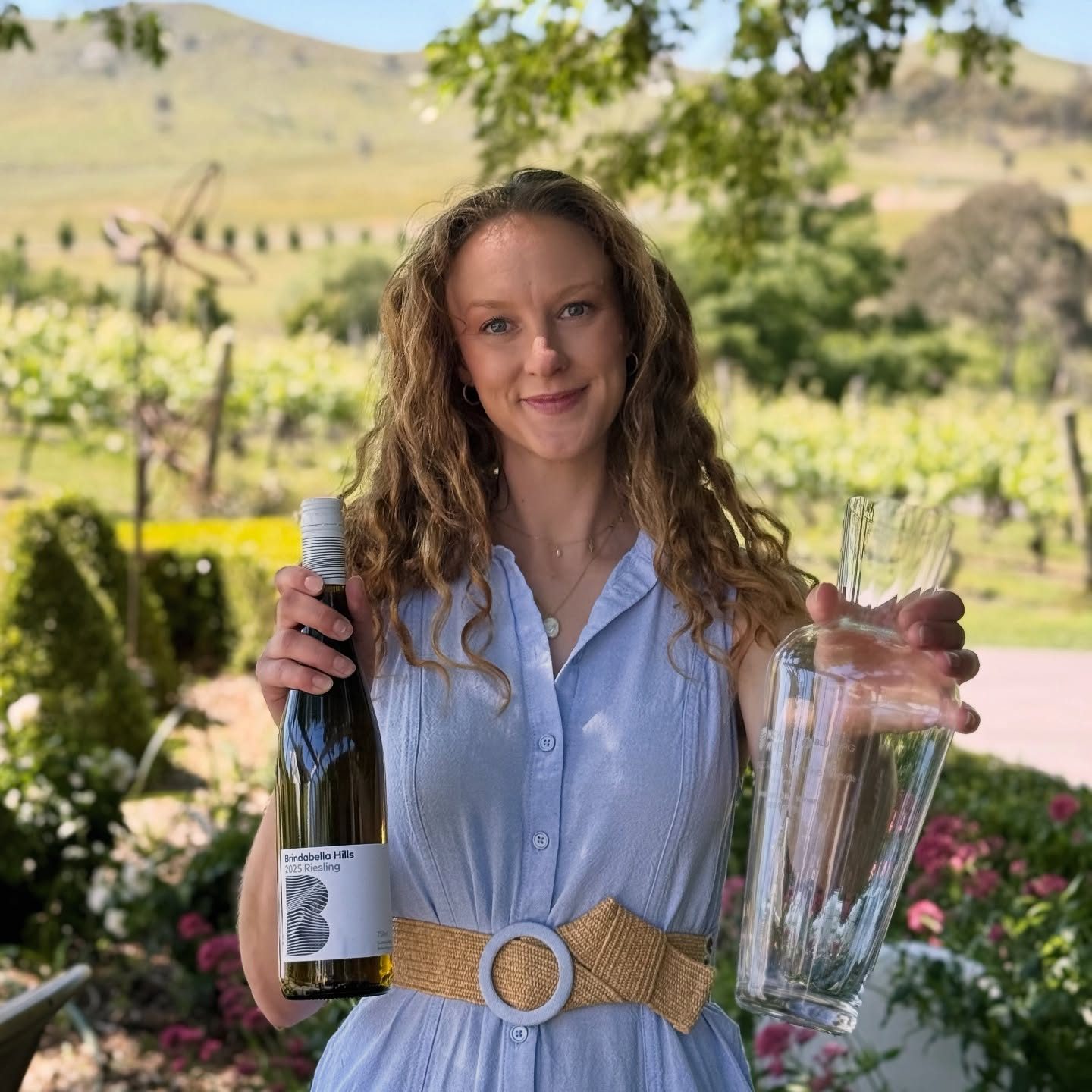 Rhiannon wears a blue button-down shirt dress with brown belt, and holds a bottle of wine in one hand and a glass trophy in another.