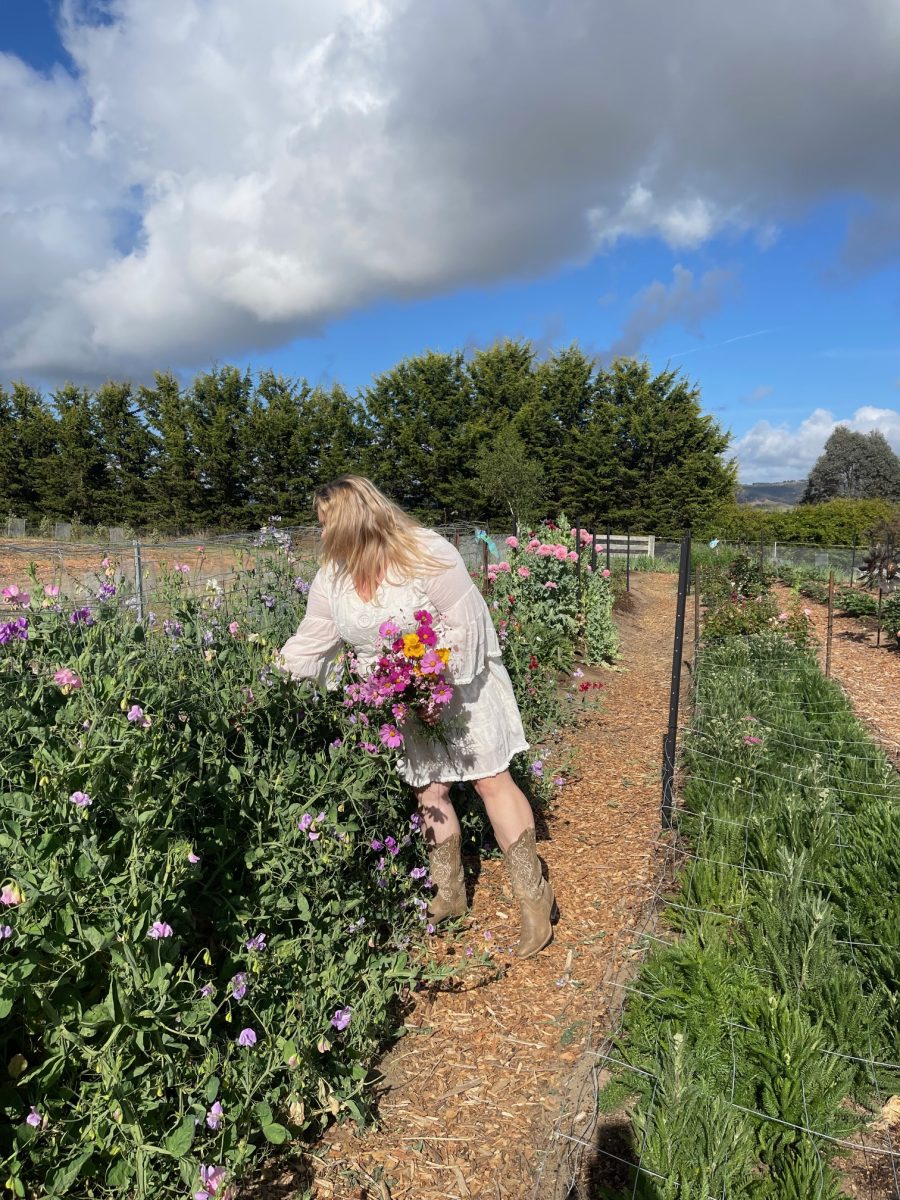 A woman in a flowing white dress and cowboy boots picks a posy of wildflowers. 