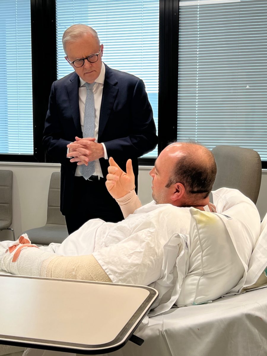 Man in suit standing by a man in hospital bed and chatting.