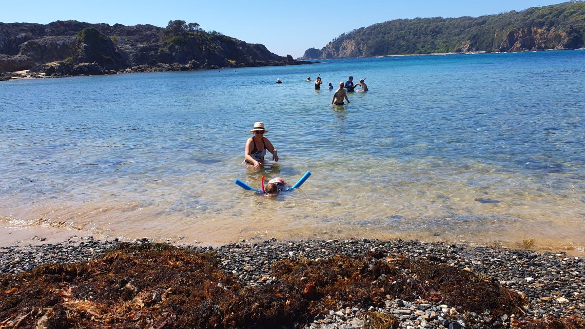 A child snorkelling, kept afloat with a pool noodle, and her mother behind her watching on - with the expansive blue waters of Guerilla Bay in the background.