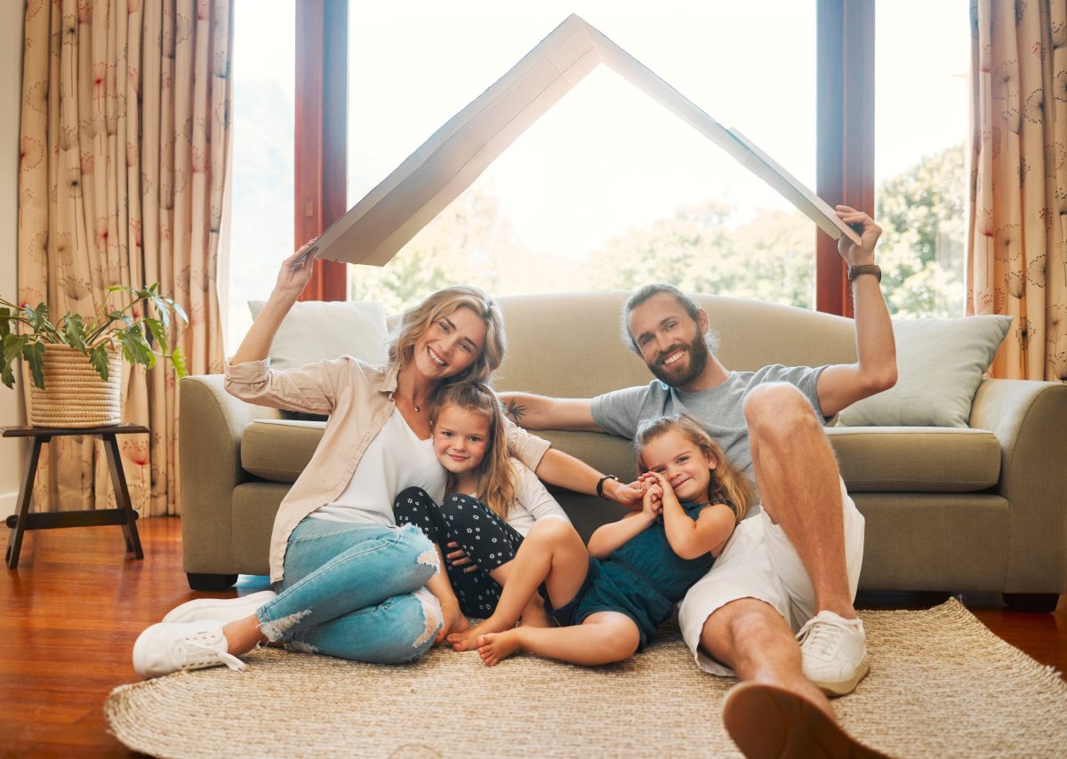 Young happy content caucasian family holding a cardboard as a roof covering them sitting on the floor