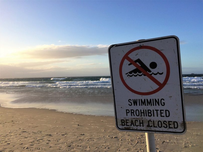 A sign on a pretty beach shows a swimmer in a circle with a red line through it and the words: 'swimming prohibited, beach closed'. The sun is setting in the distance and the day is about to end. The photo was taken in Bryon Bay, Australia.