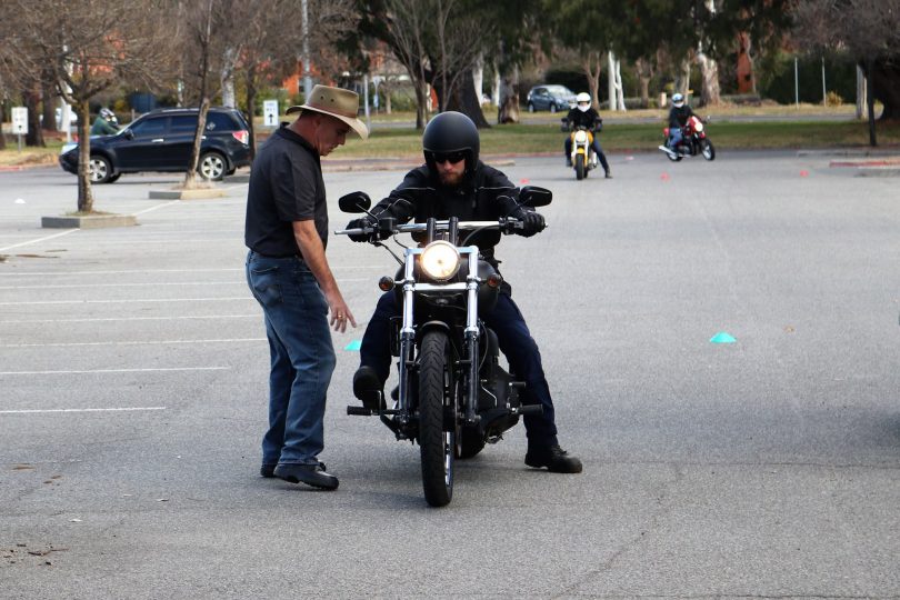 Instructor standing next to a rider on a motorcycle, pointing towards the front wheel as he talks to the rider