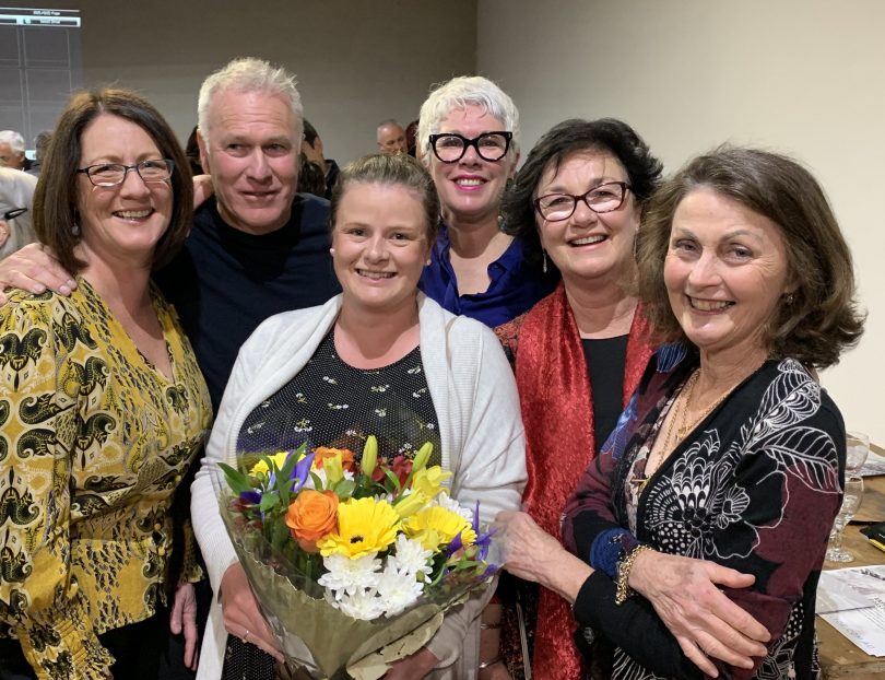 Six people gathered together and smiling at the camera, with the woman in the middle holding a bouquet of flowers