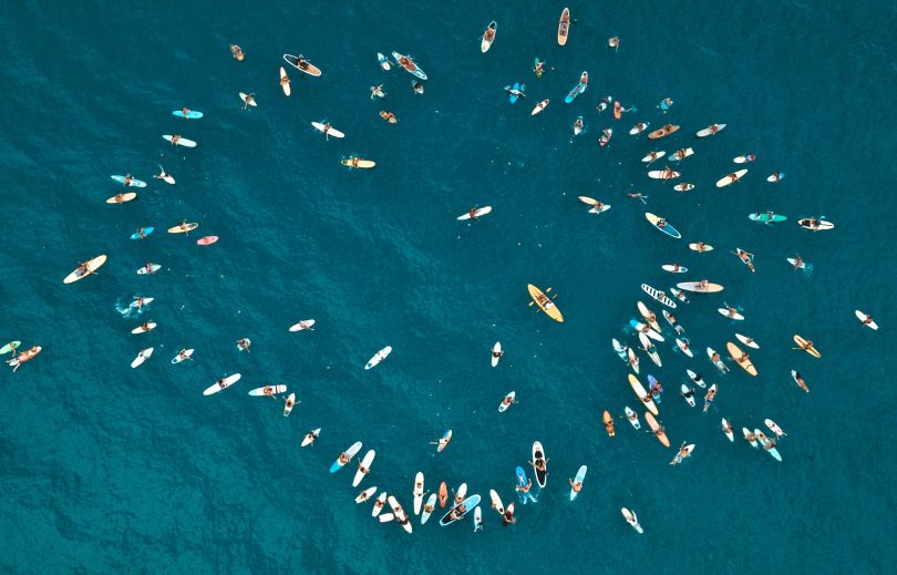 Aerial view of paddle-out gathering on paddle boards in open water.