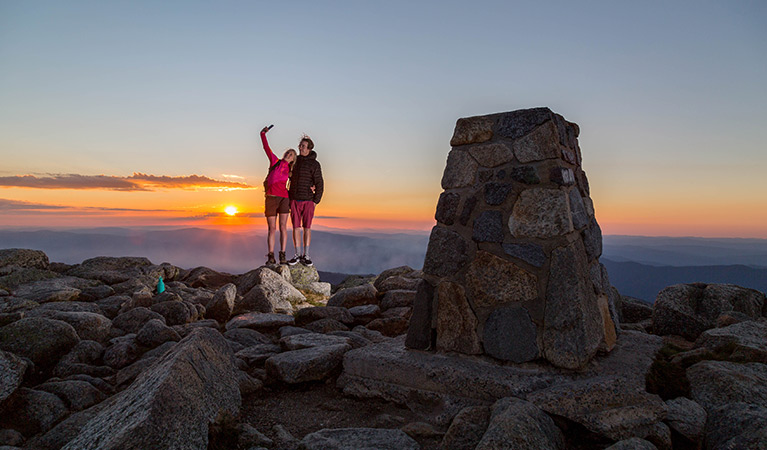 Man and woman stand at summit of Kosciuszko National Park with sunset behind them.