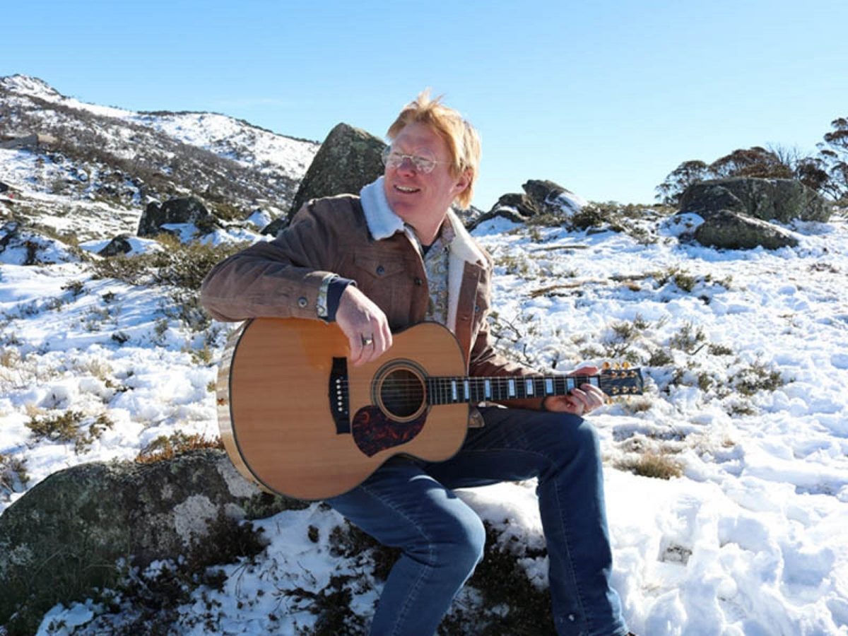 Darren Coggan perched on a rock in the snow and holding his guitar
