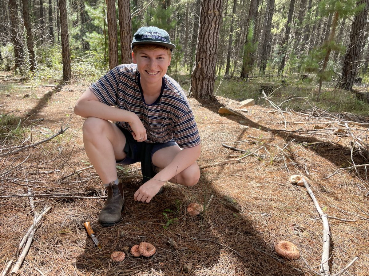 Marita, wearing striped t-shirt, shorts and a cap that says 'happy shrooming' crouches in a forest near a group of orange mushrooms.