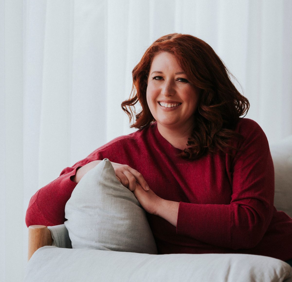 Jane Harper in a red shirt and smiling at the camera while leaning on the back of a couch