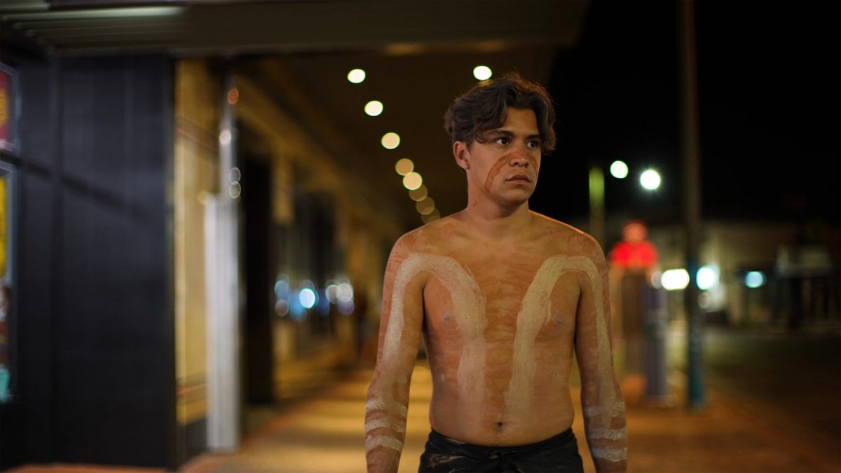 A shirtless young man with white and brown paint on his torso and face at night