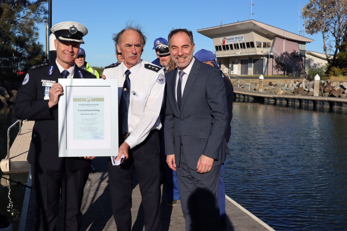 Three men standing beside a river, with the two on the left holding a framed piece of paper