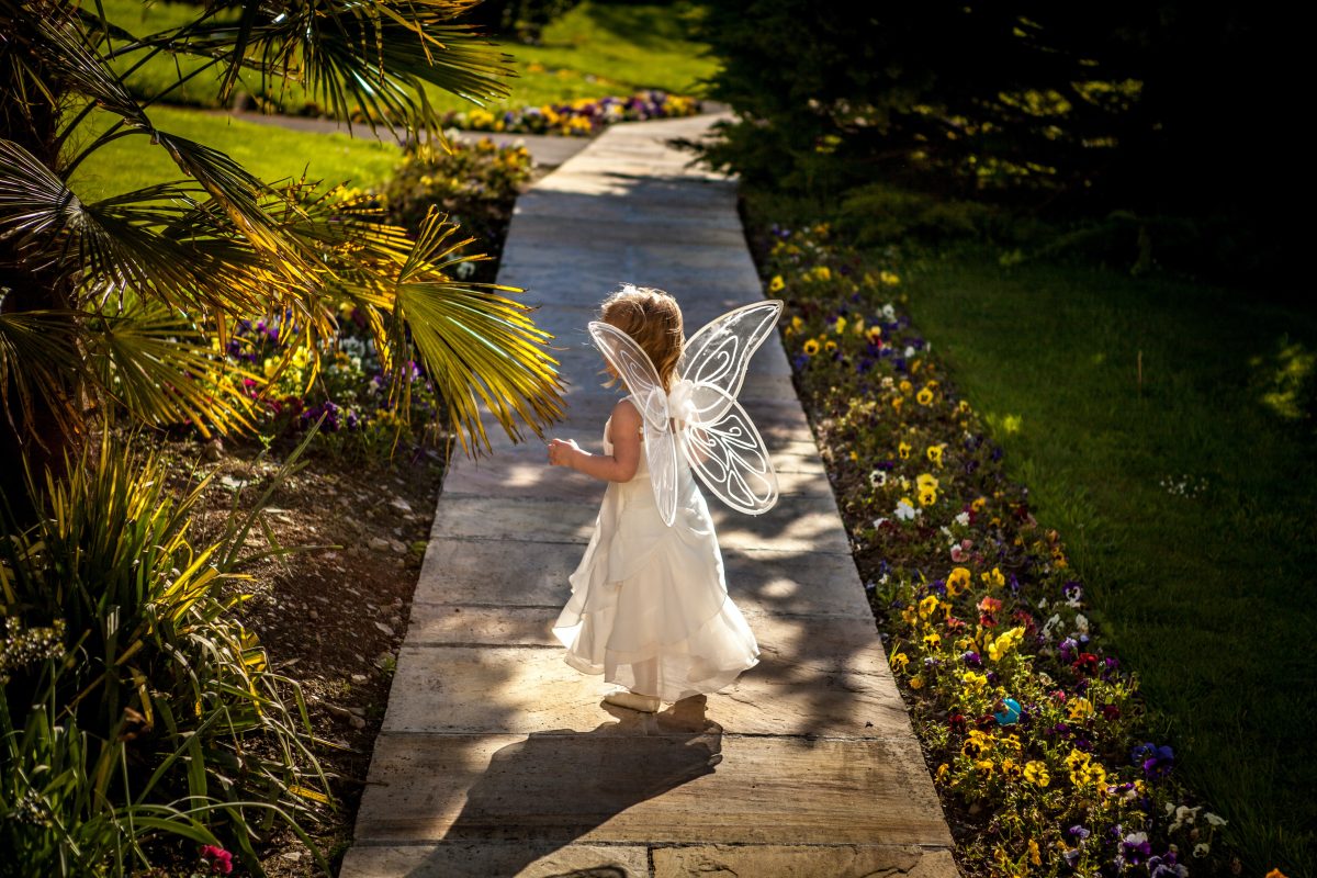 A young girl turned away from the camera in fairy wings, walking down a pathway