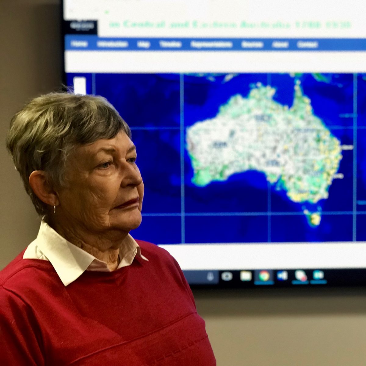 Professor Lyndall Ryan in a red jumper standing in front of a screen with a map of Australia on it.