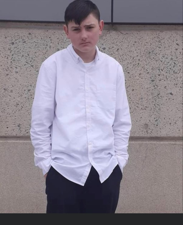 A photograph of a young boy in a white shirt and black pants looking at the camera 