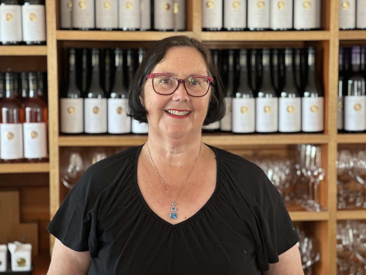 A White woman with short black hair and red glasses in front of a shelf of wines