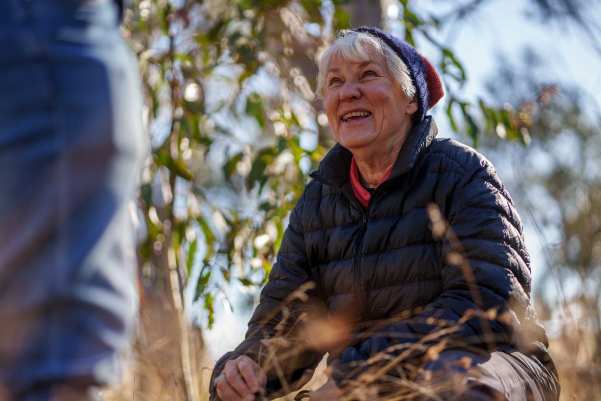 Barbara Evison in a parka and beanie crouching over some grass while looking up and smiling at the person standing before her.