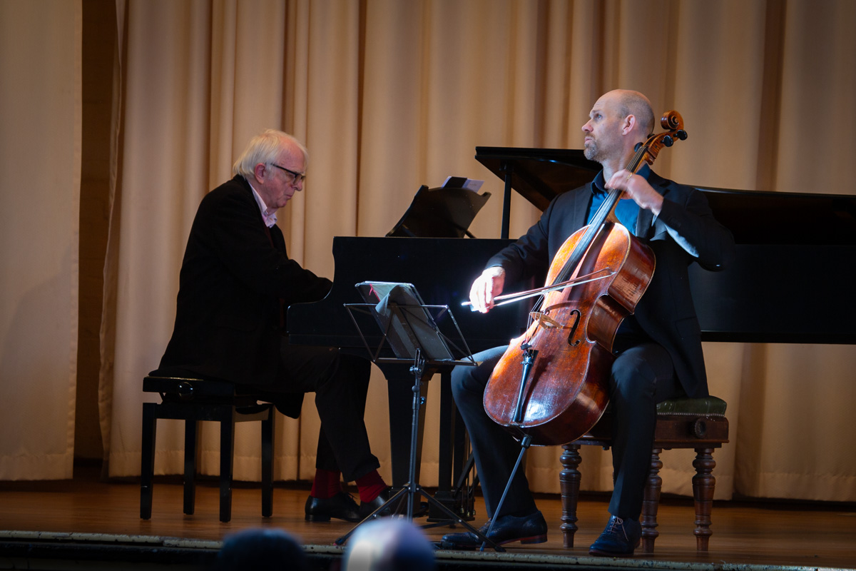 Two musicians on stage performing to a crowd. One in the background playing a grand piano, the other in front playing cello.