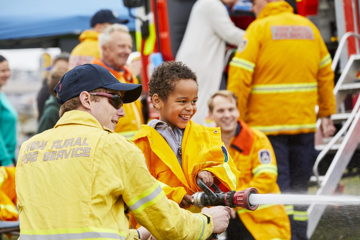 Adults in orange NSW RFS uniforms and a young boy aiming a hose
