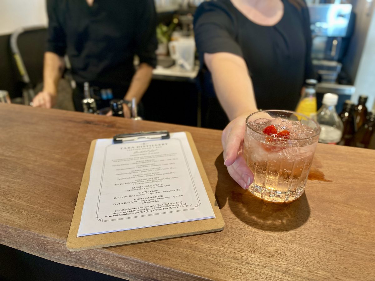 A bartender hands a glass of gin and tonic, garnished with a strawberry, across the bar