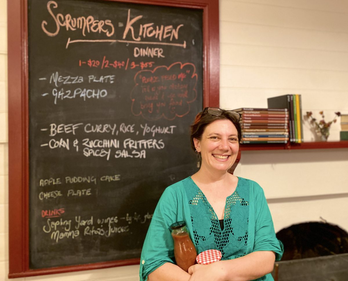 Ruth holds preserves in bottles and stands in front of blackboard menu reading Scrumpers Kitchen.