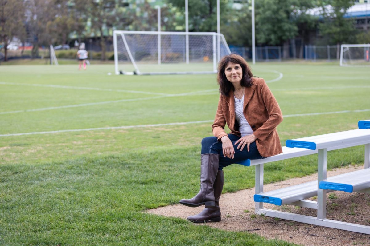 A woman in a brown coat and jeans sitting on a bench next to a sports field