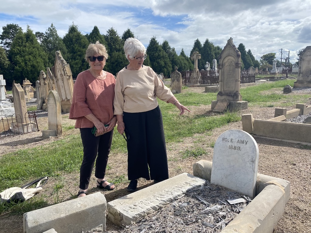 Two women at a graveyard, one pointing at a specific grave
