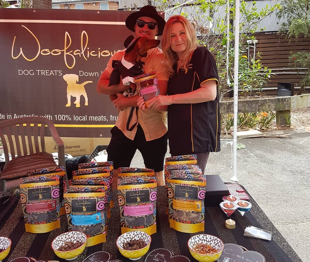 Two people standing and holding a dog in front of a table of dog treats