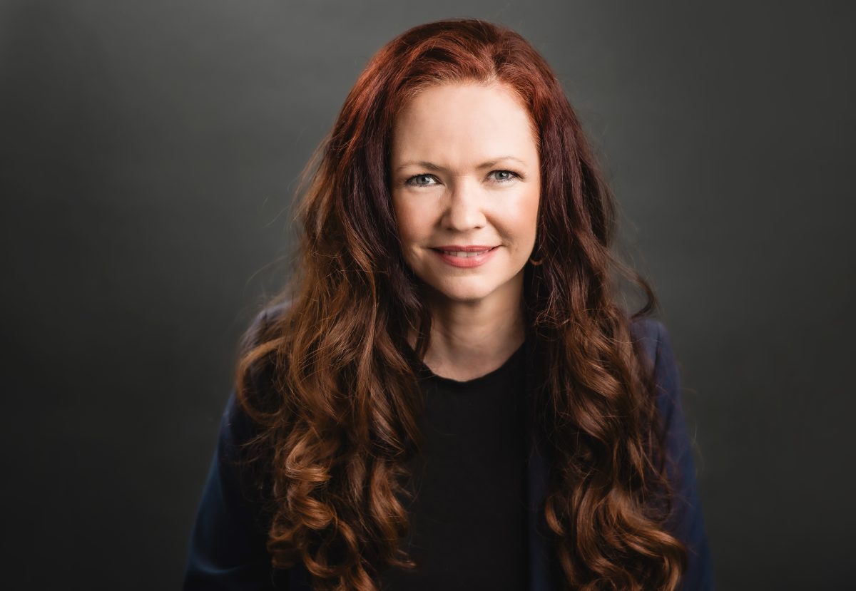 A headshot of a woman with long red hair looking straight at the camera