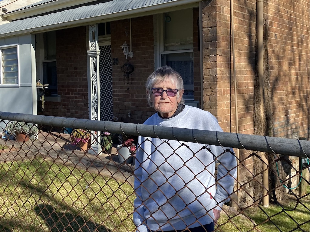 elderly woman standing at the front fence of her house