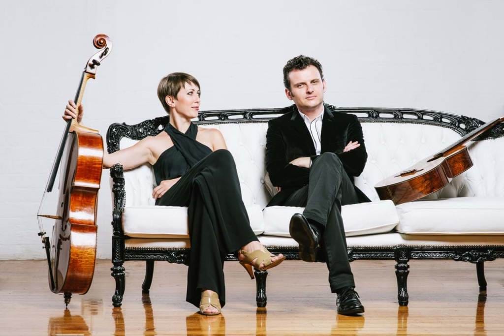 Two people in black clothes sitting on a white couch with musical instruments