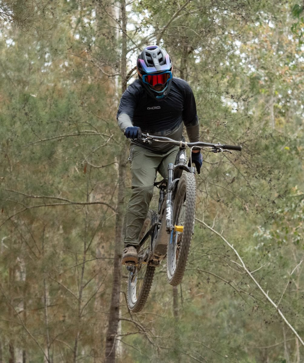 Mountain biker takes a jump with one hand off the bars.