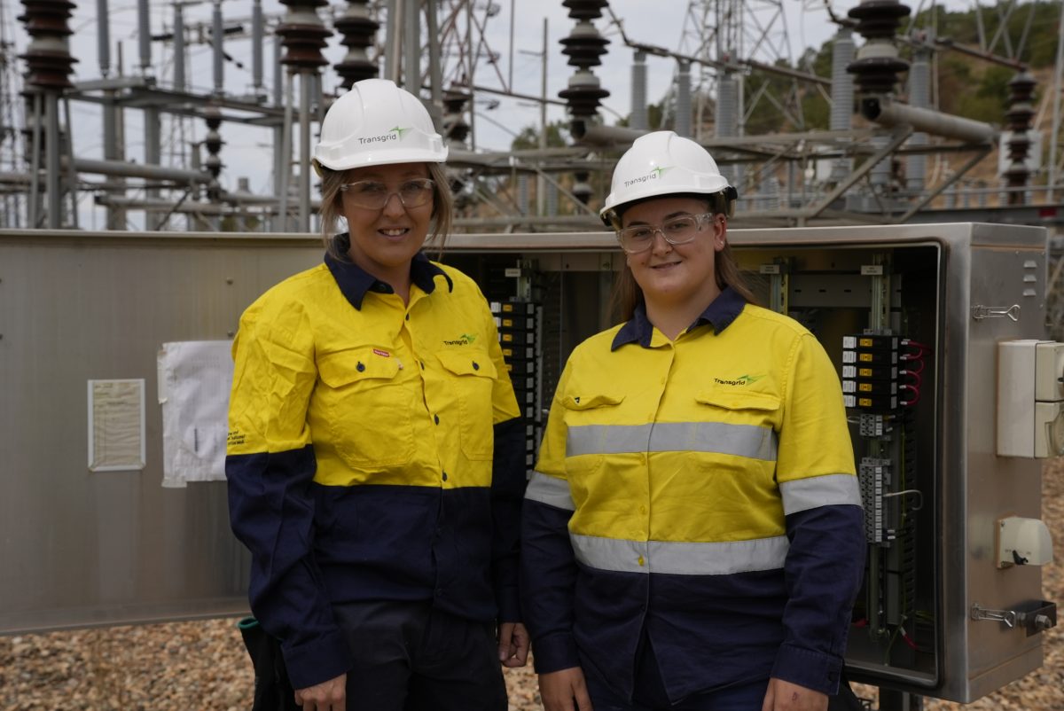 Two women in hard hats and in yellow fluro in front of power grid.