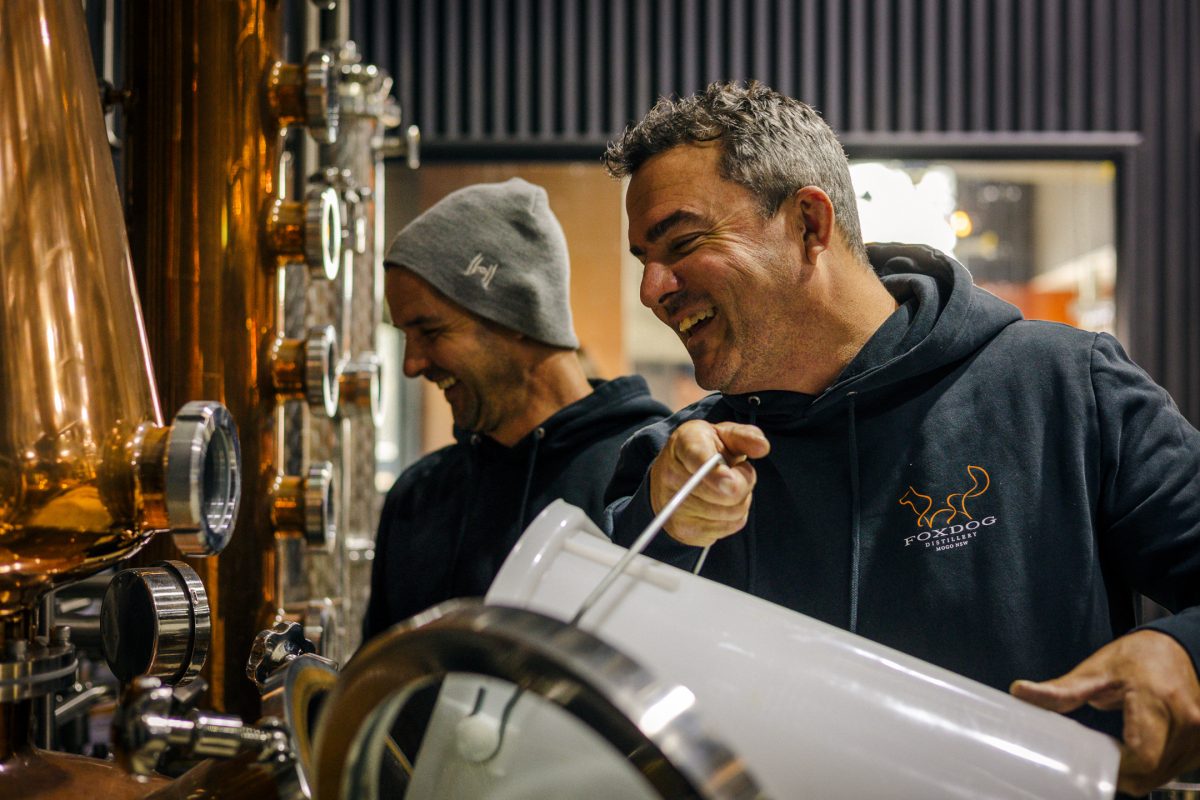 Two men smile while pouring liquid from a white bucket into a copper still