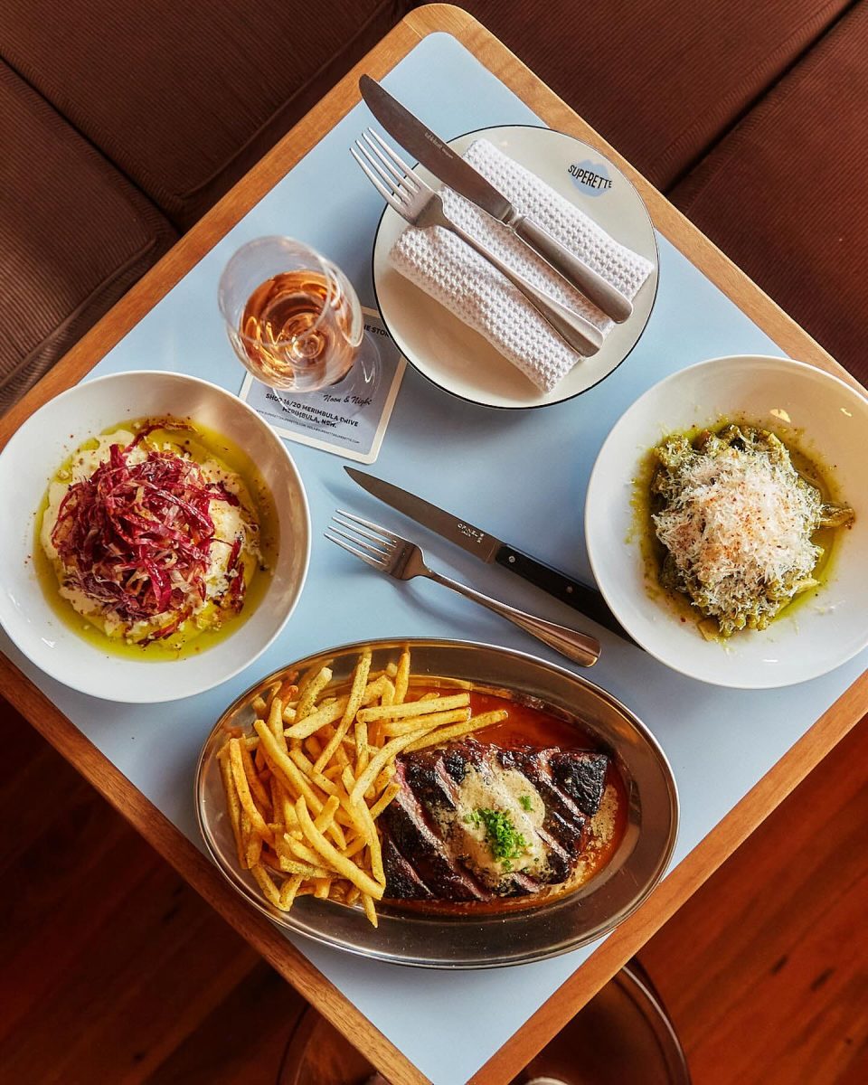 A table with a plate of steak and fries, two side dishes and a glass of wine