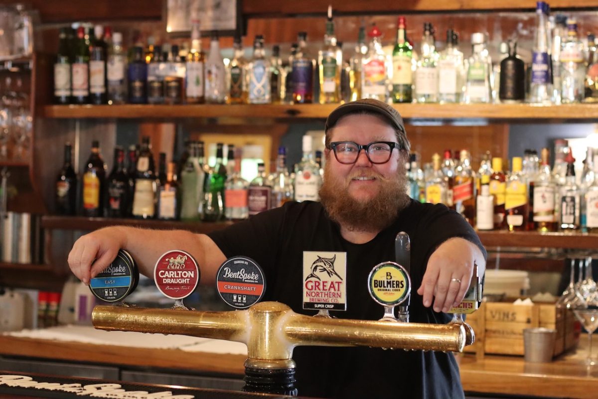 A bearded man in glasses rests his arms on beer taps.