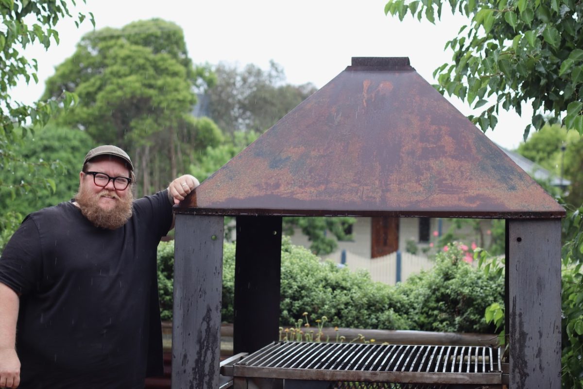 A man in a beard leans against a large outdoor barbecue.