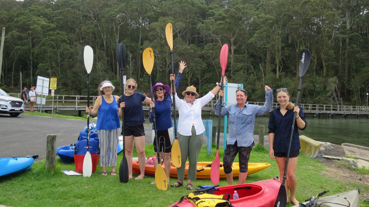 A group of women standing near kayaks and holding paddles