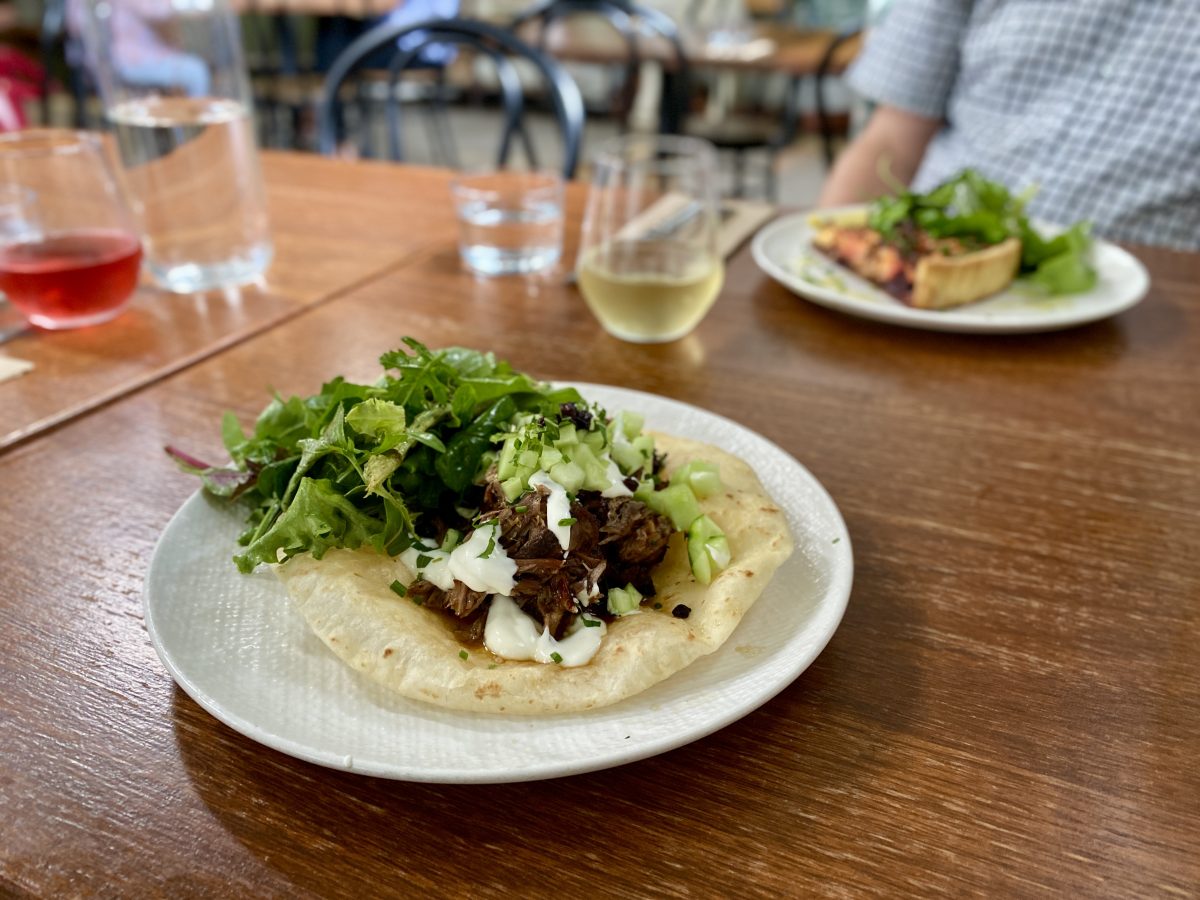 A plate with flatbread piled with pulled meat, with salad and chopped cucumber, a glass of wine and a tart is in the background