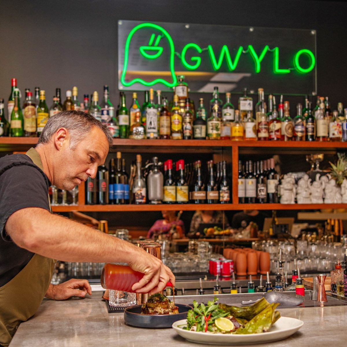 A chef adds sauce to a dish, with a green neon sign reading GWYLO in the background above a bar