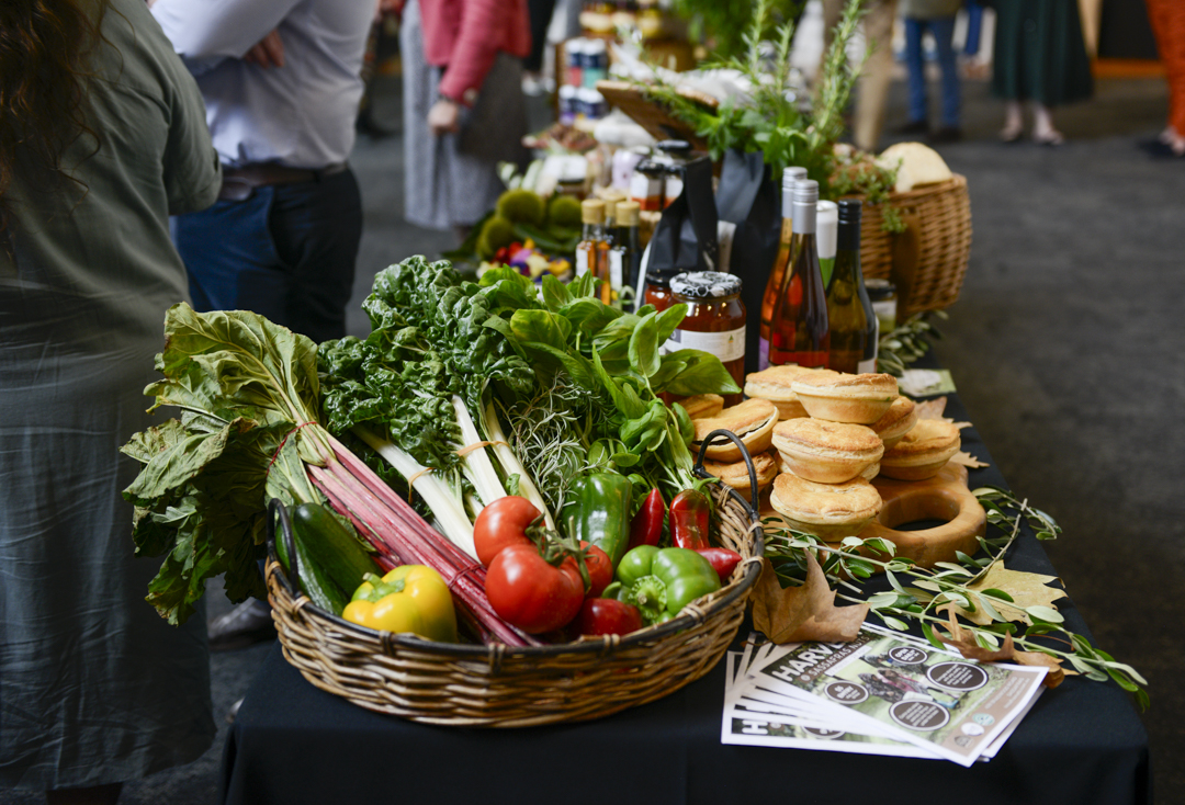 A basket of fresh fruit and vegetables on a table laden with baked goods, wines and other foodie products